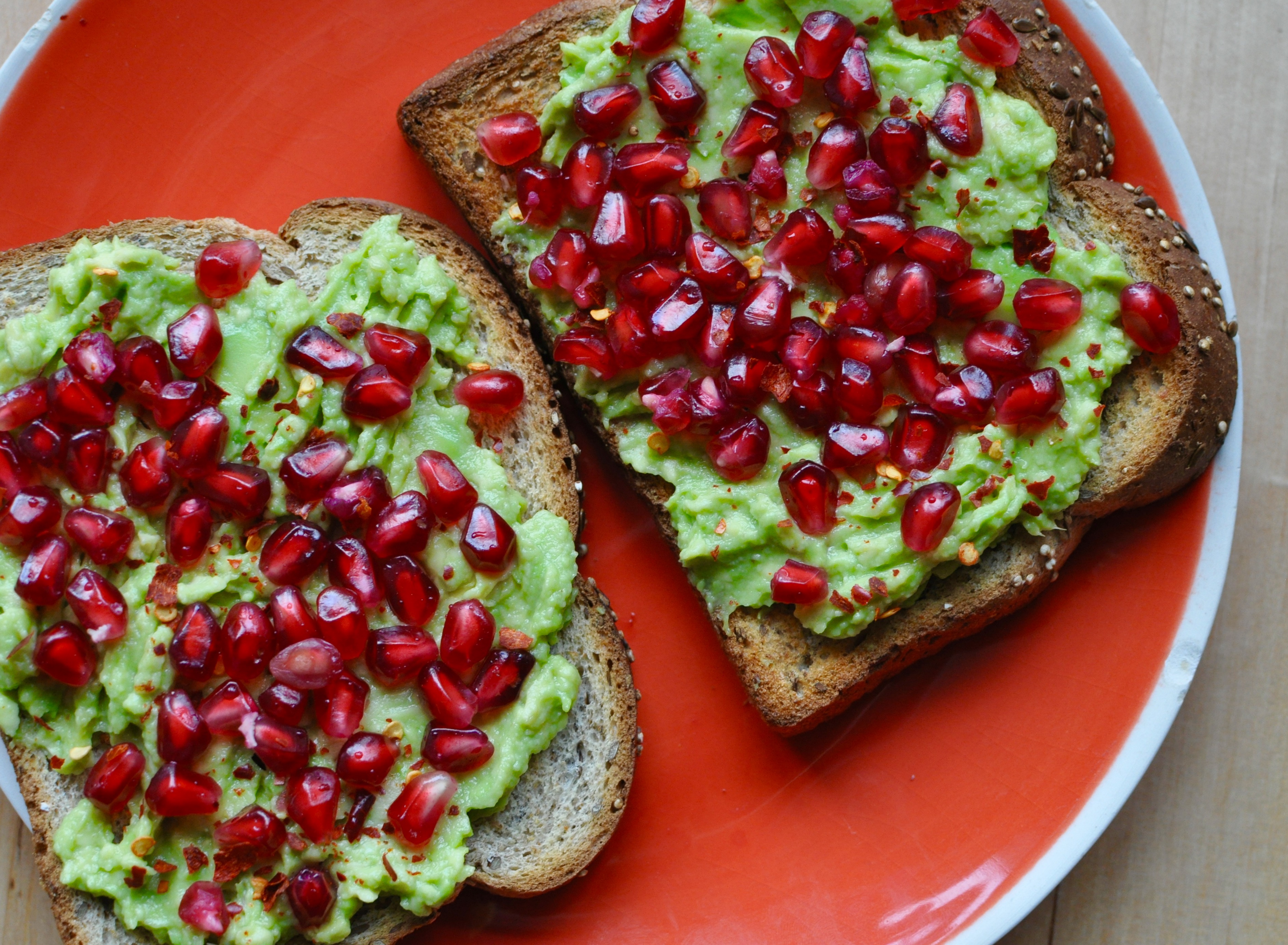 Toast on an orange plate topped with avocado, pomegranate seeds, and red chillies.