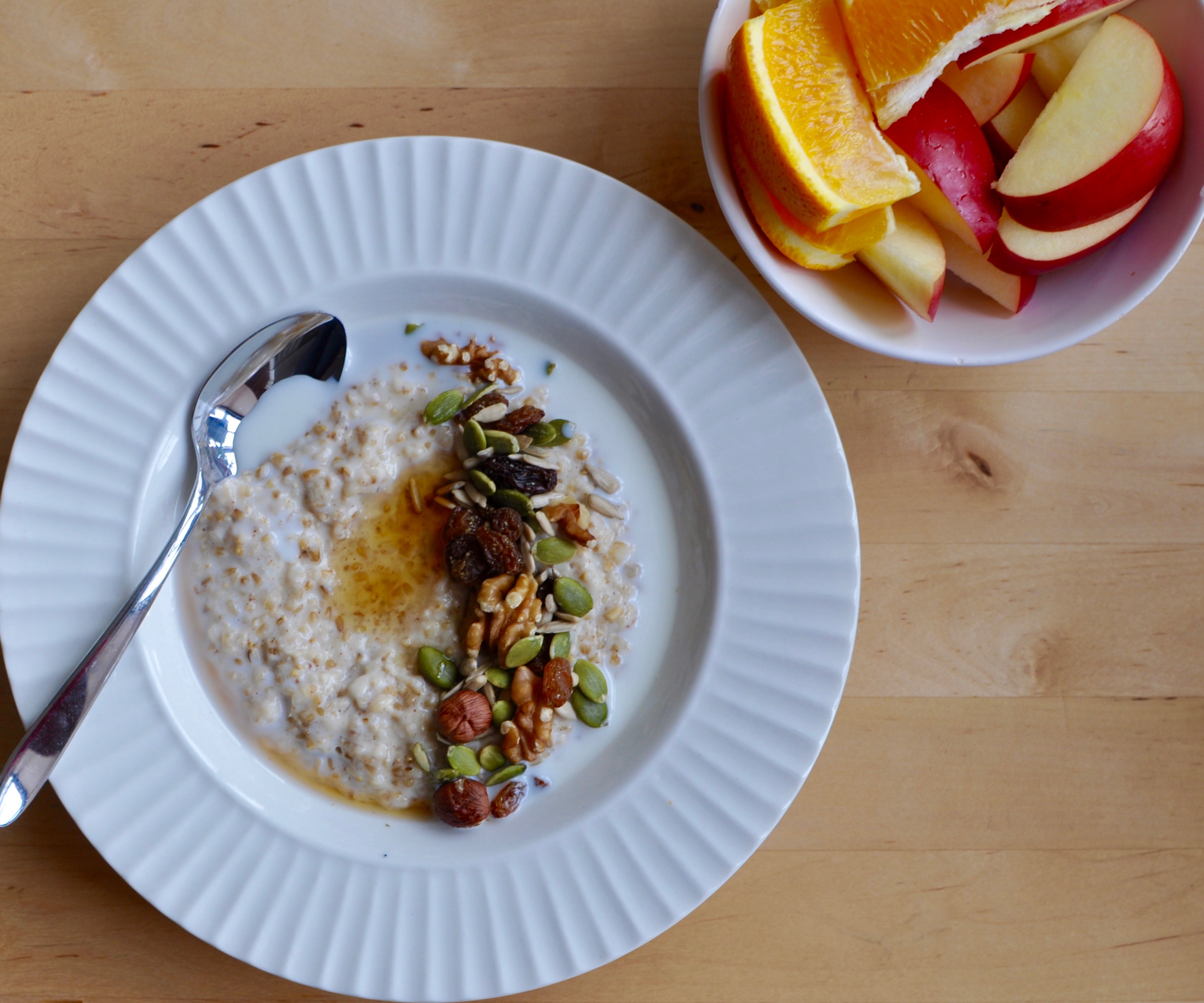 A shallow bowl full of steel cut oats, nuts, seeds, and brown sugar beside a bowl of orange sections and apple slices.
