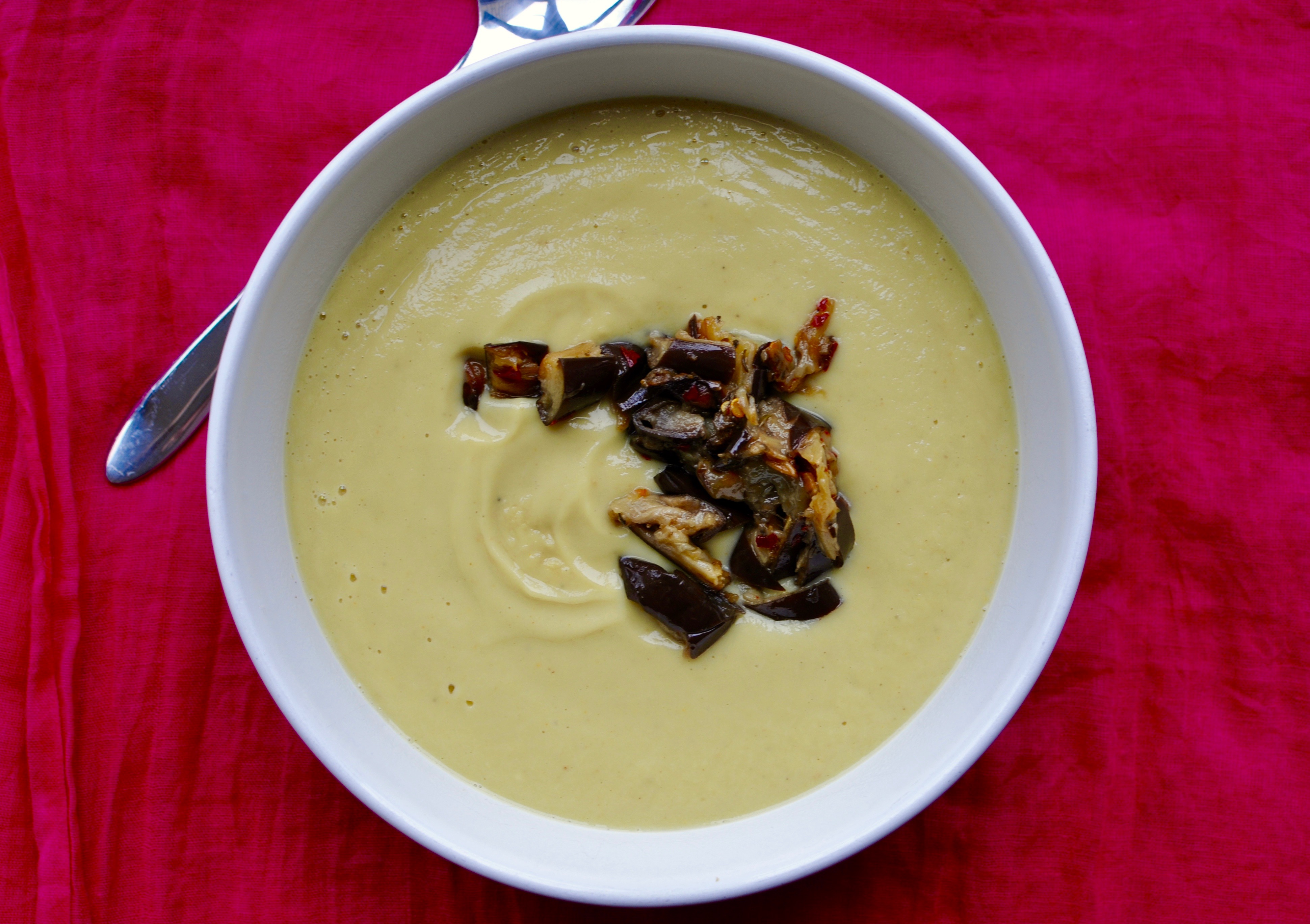 White bowl of creamy parsnip soup with eggplant as a garnish on a hot pink background.