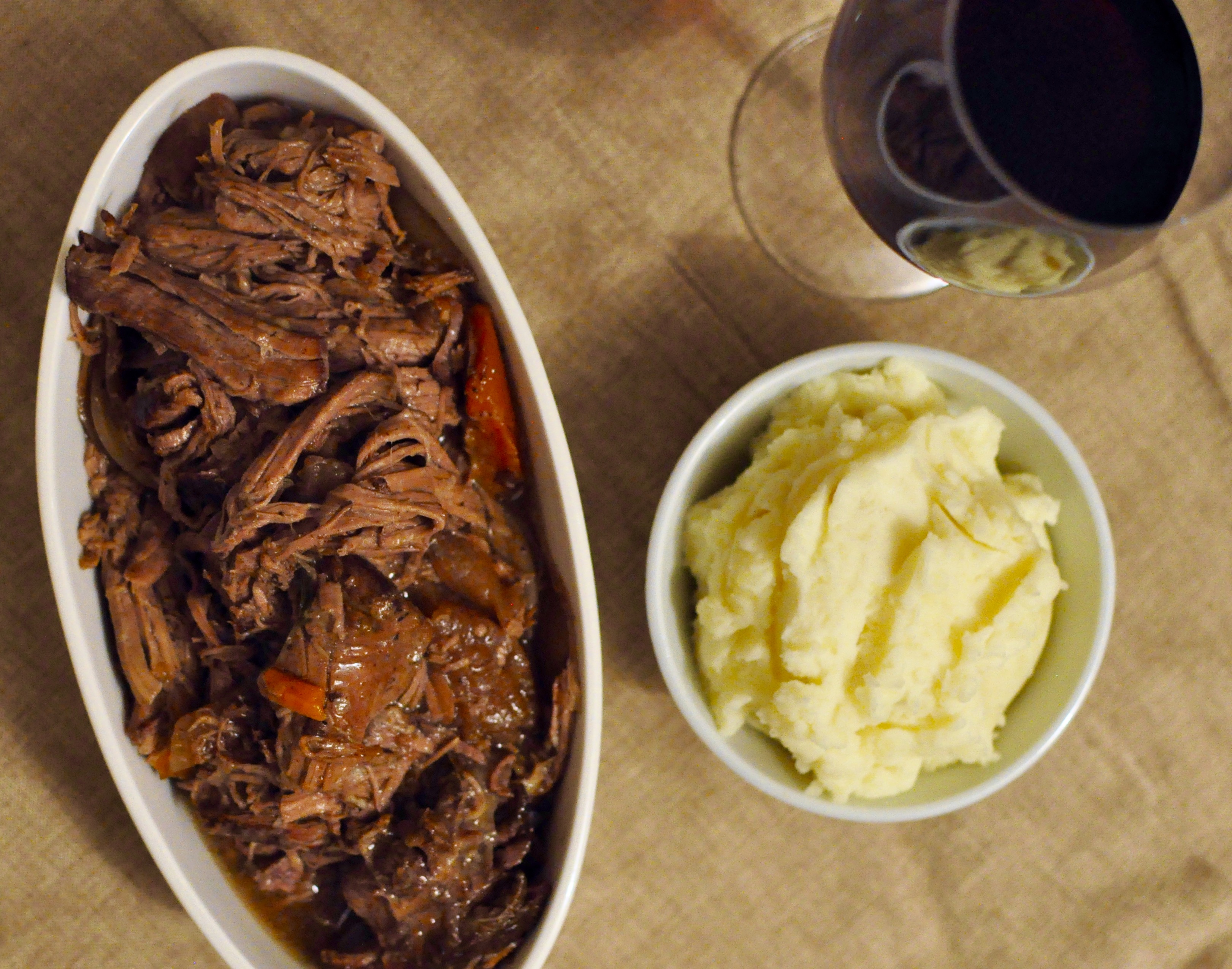 Pot roast in a narrow serving vessel with a glass of red wine and a small white bowl of mashed potatoes.