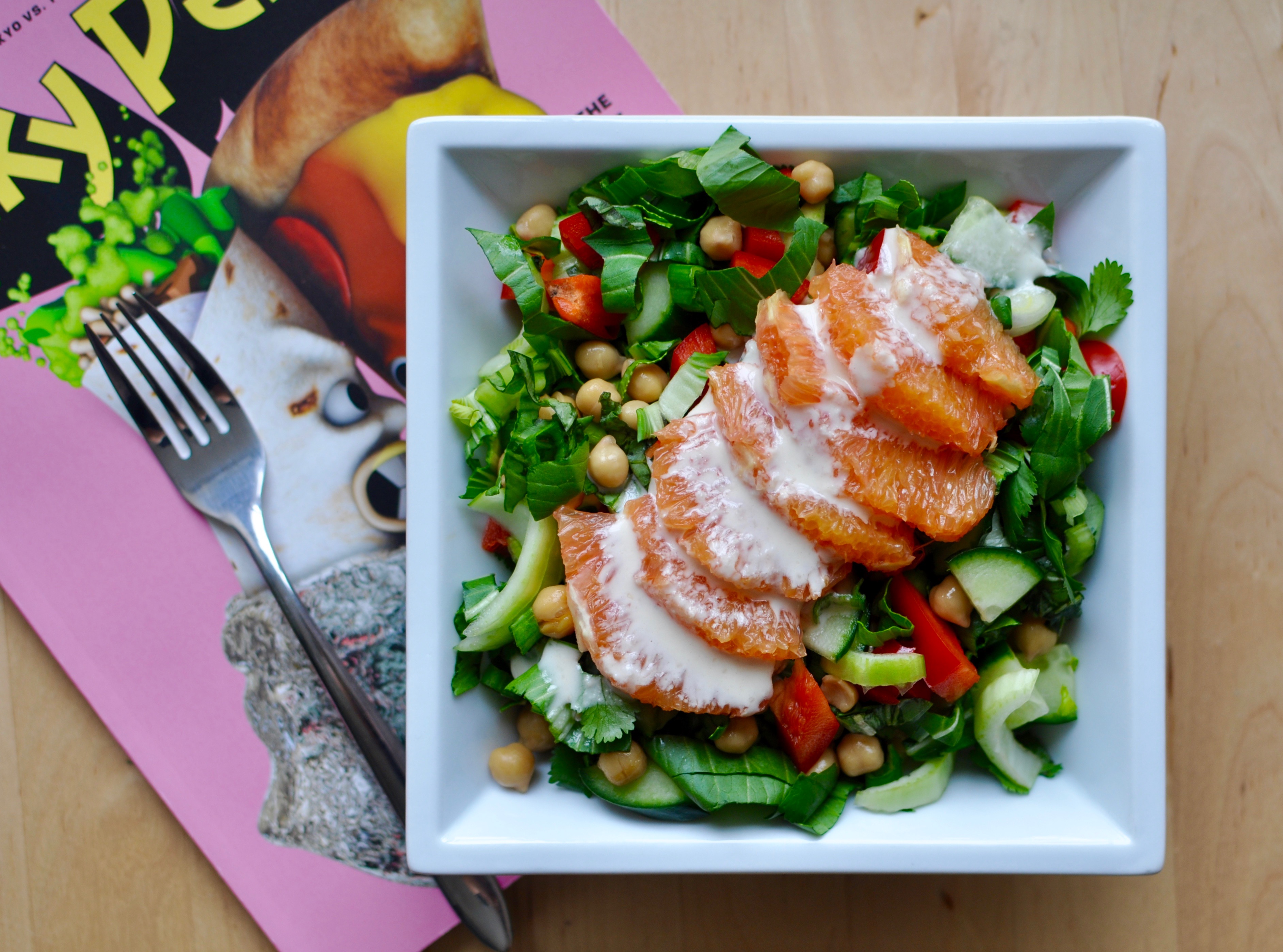 A white square plate full of salad containing chickpeas, bok choy, red peppers, and orange slices covered with a tahini orange dressing. A copy of Lucky Peach is in the background.
