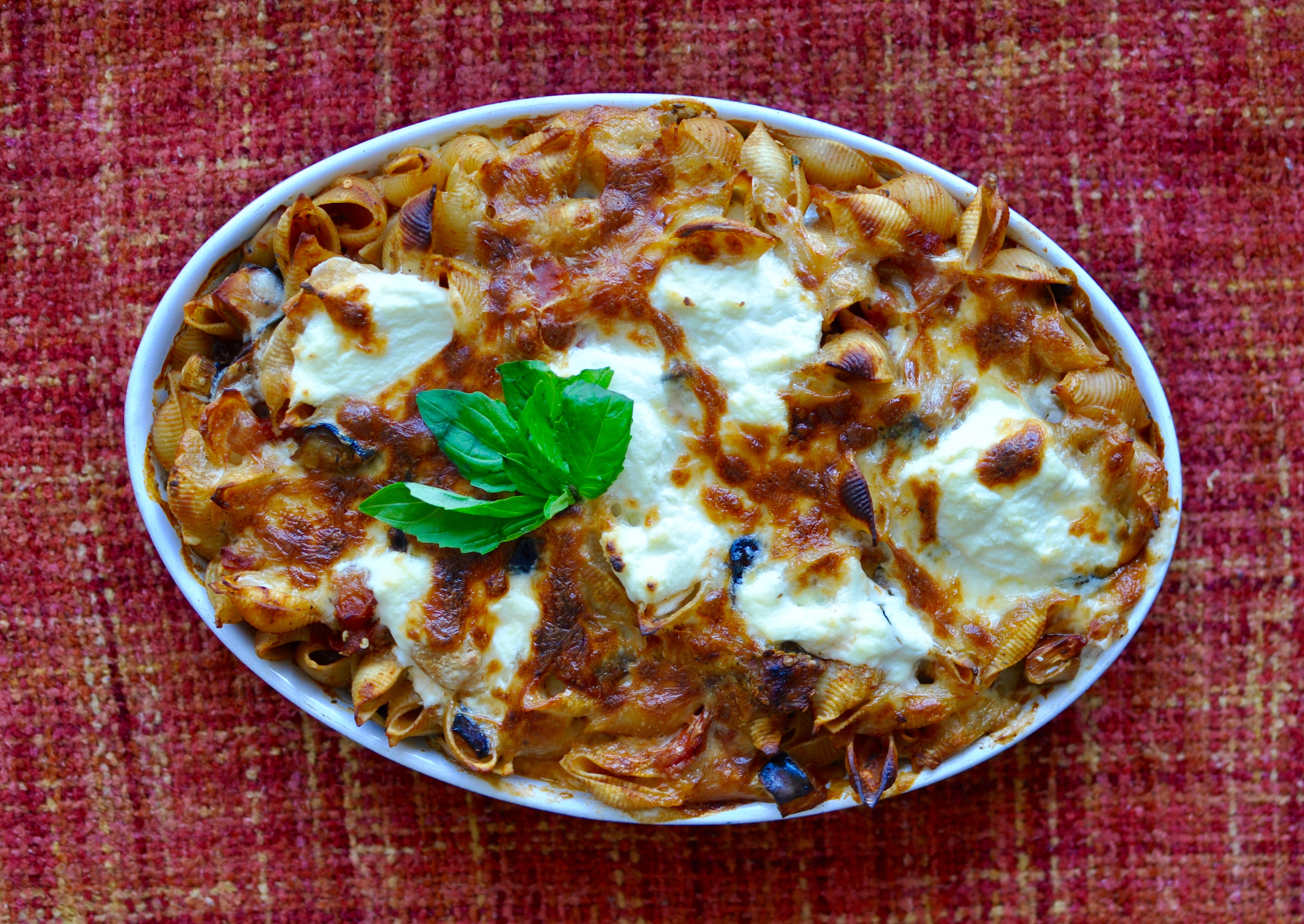 Blue oval baking dish full of pasta shells, tomatoes, eggplant, and ricotta cheese on an orange background.