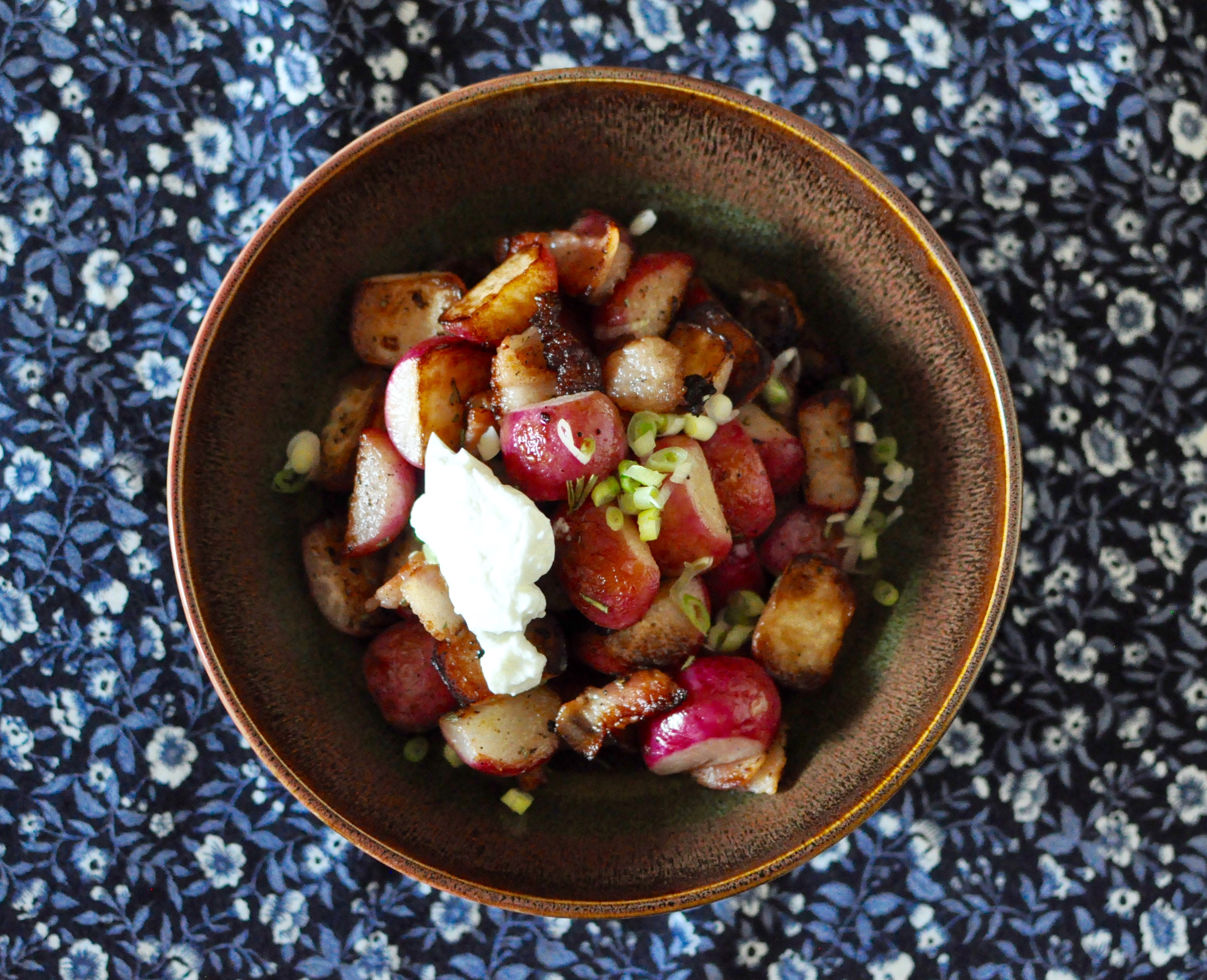 A brown shallow bowl full of roasted radishes, bacon, scallions and a small tablespoon of skyr on top.