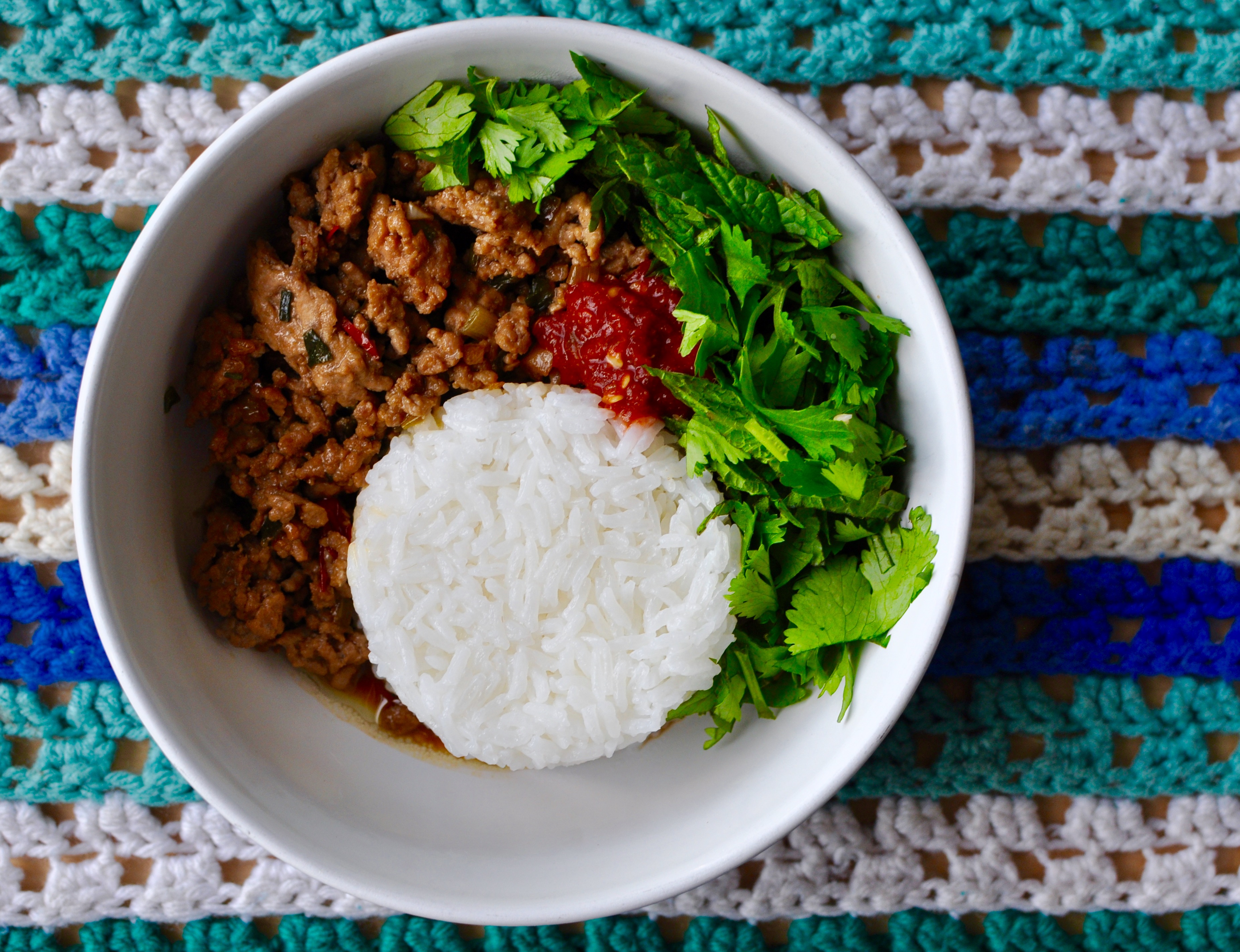 Round white bowl with white rice, minced pork, chili sauce, and fresh herbs.