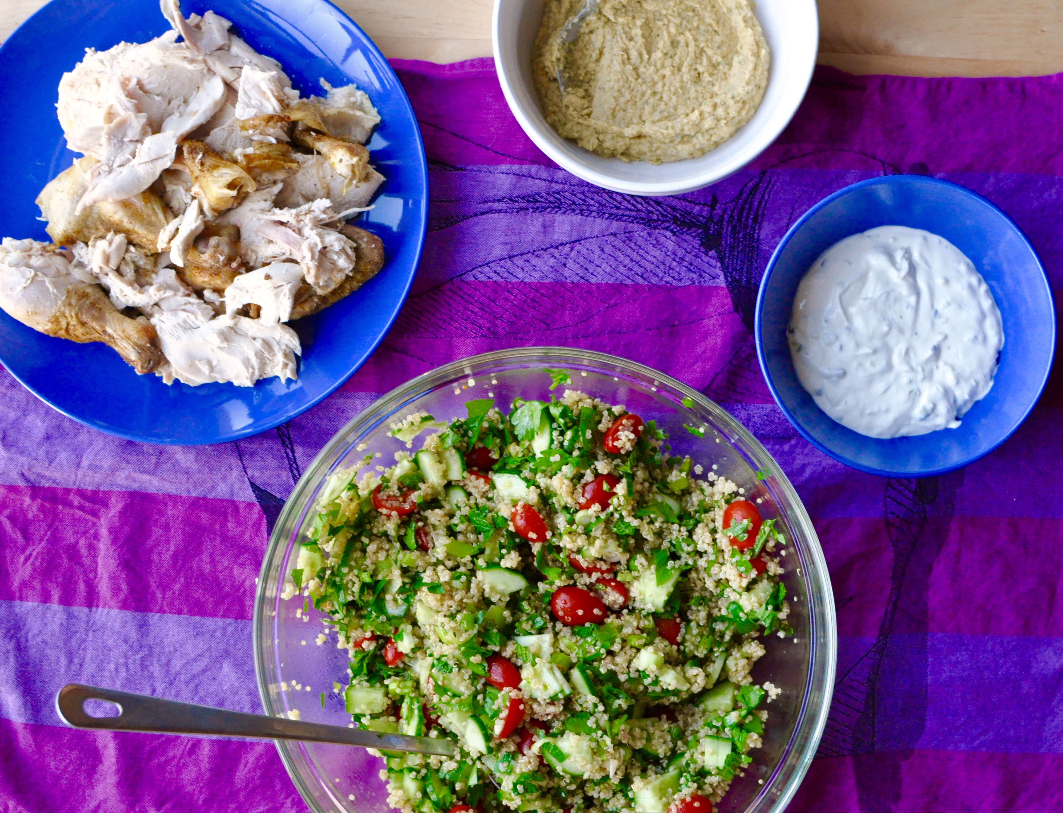 Roast chicken, tabouleh, hummus, and tzatziki arranged in serving dishes on a bright purple tablecloth