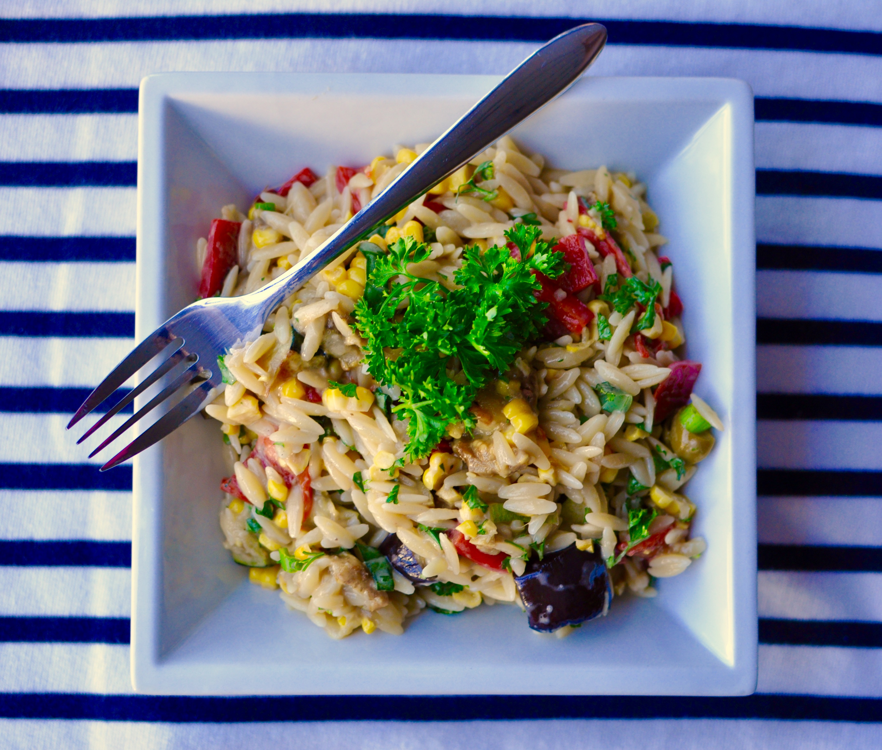 Square white bowl with creamy orzo pasta salad with roasted red peppers, zucchini, and eggplant. On a blue and white striped tablecloth.