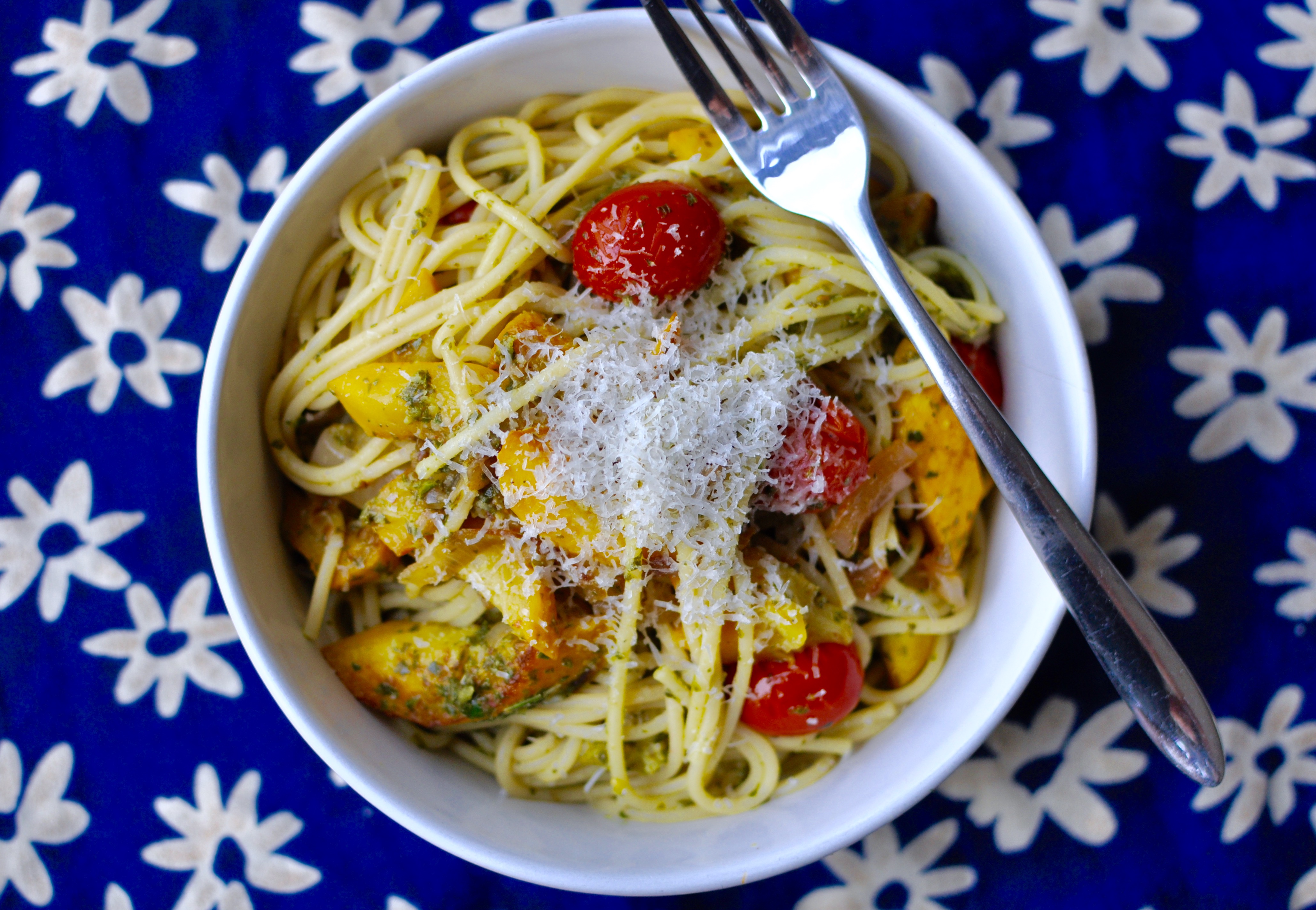 A round white bowl full of spaghetti, yellow squash, and grape tomatoes covered with green pesto and topped with parmesan cheese,.