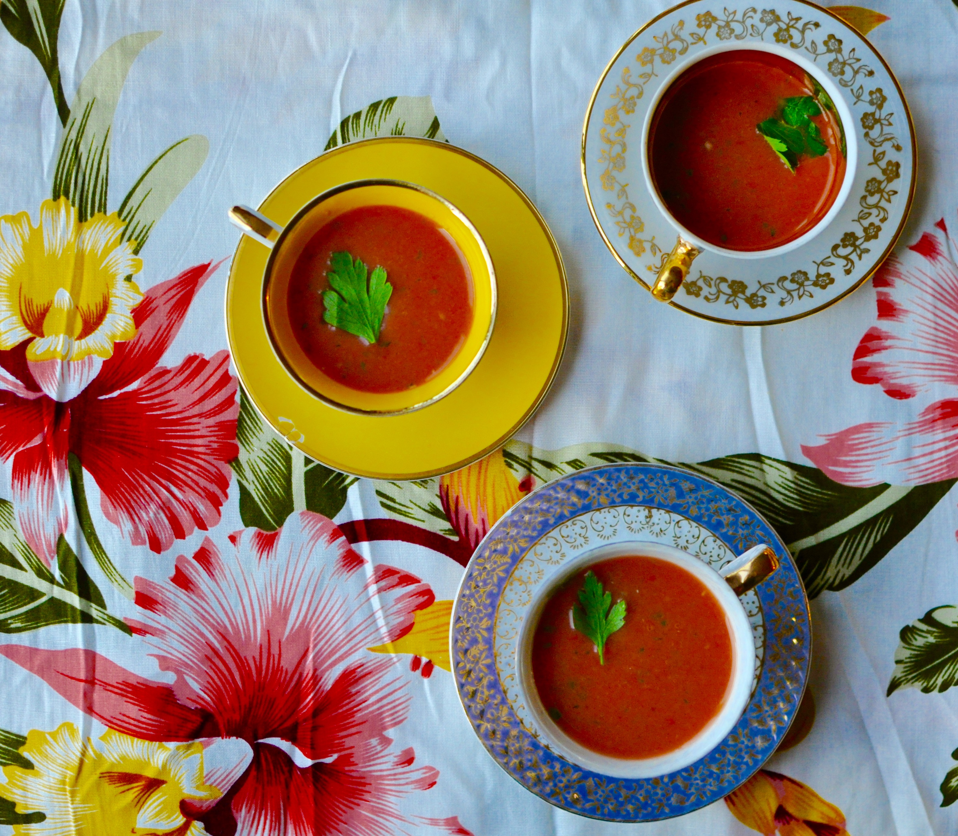 Colourful teacups on a flowered tablecloth, filled with pink watermelon gazpacho.