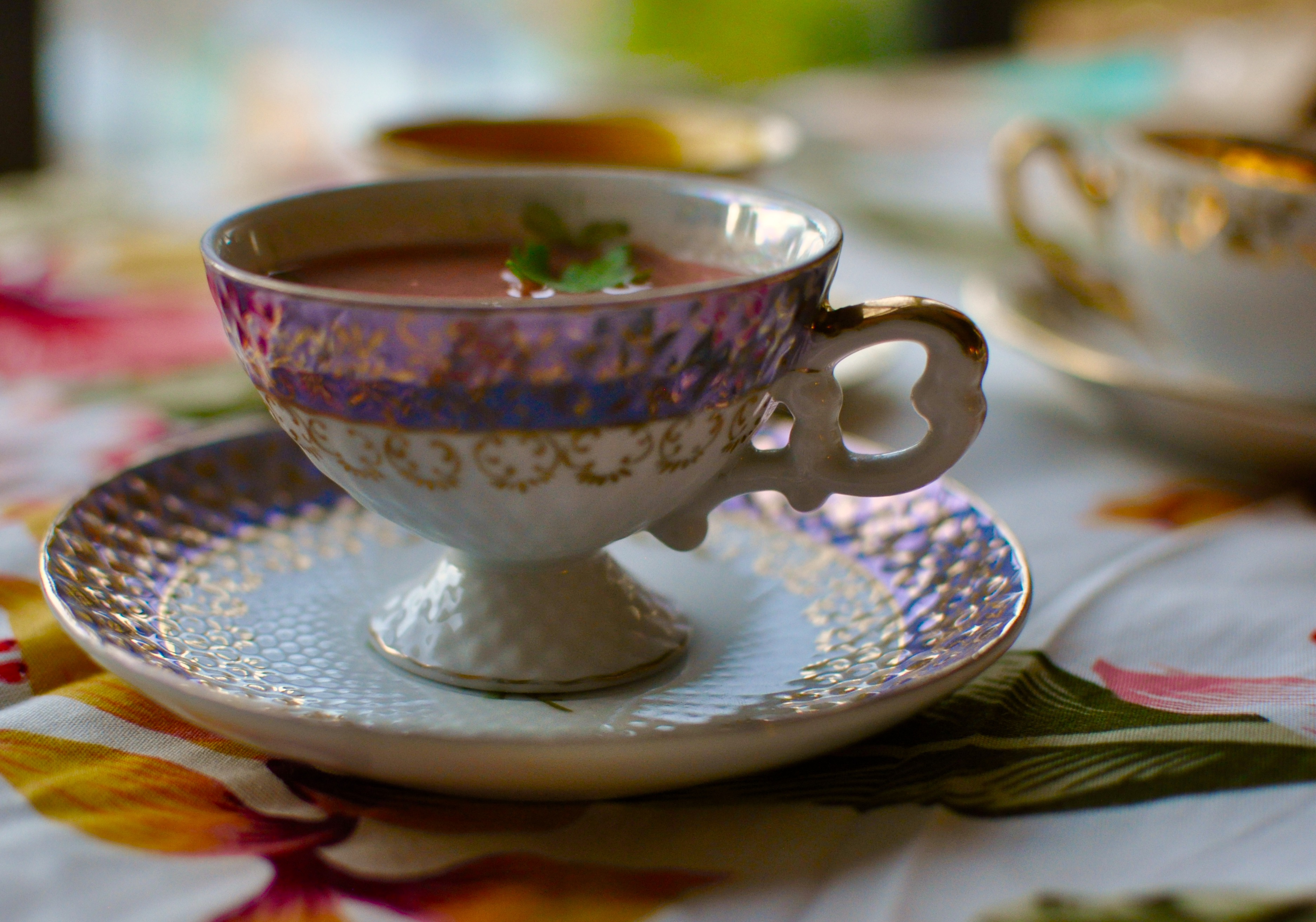 Colourful teacups on a flowered tablecloth, filled with pink watermelon gazpacho.