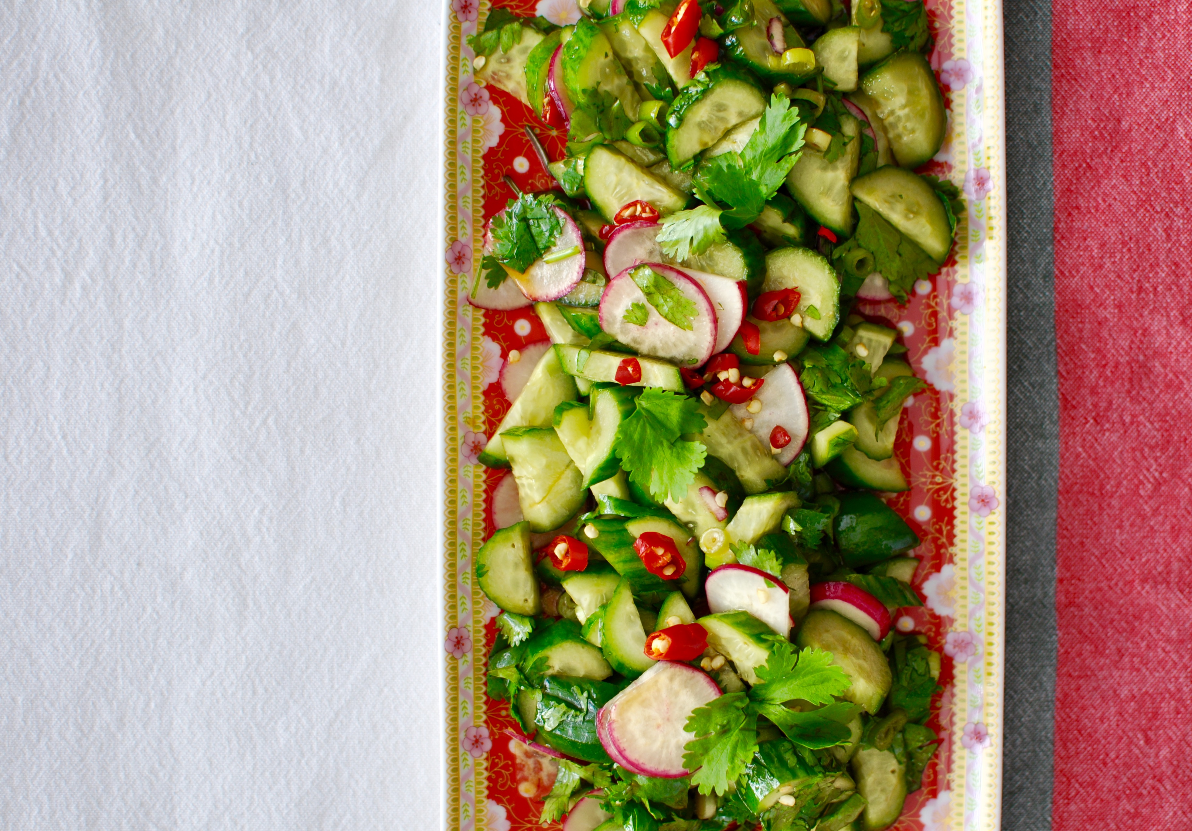 Rectangular plate with cucumber, radish, and chilies garnished with cilantro. On a white, slate, and red tablecloth.