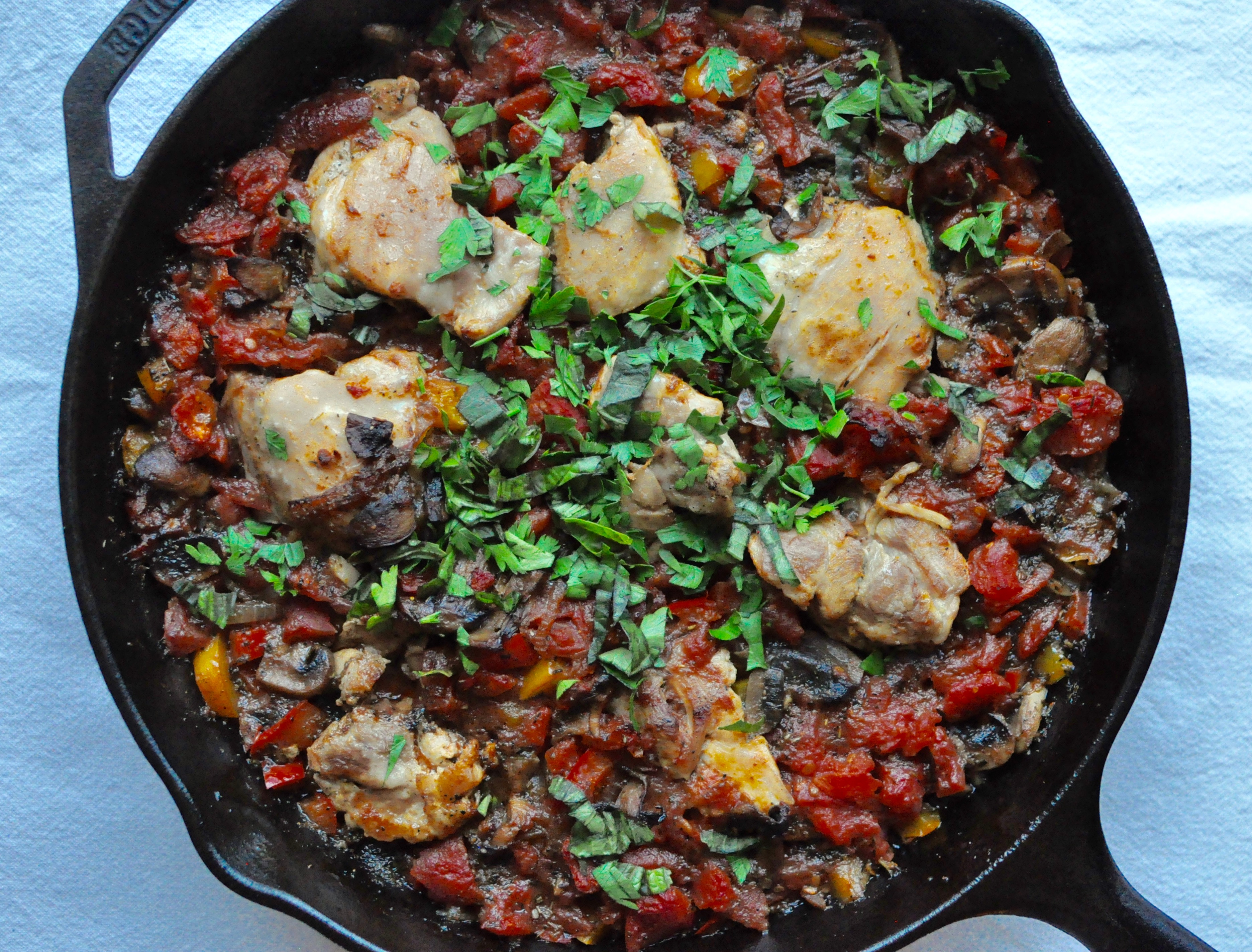 Large cast iron skillet full of chicken cacciatore sprinkled with fresh herbs on a blue tablecloth.