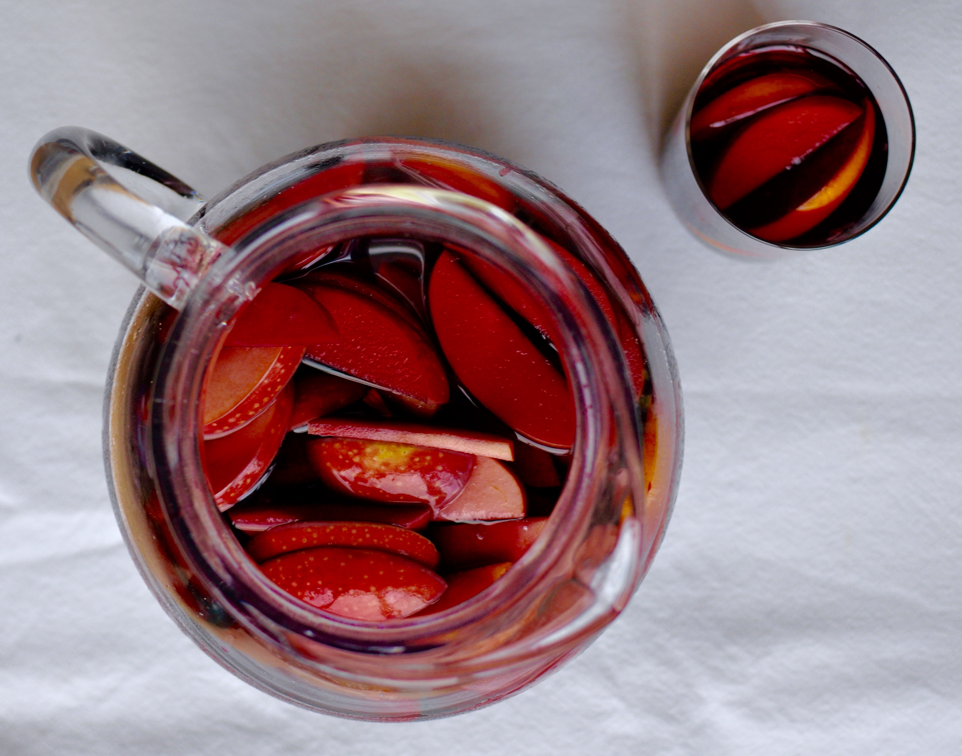 Classic red wine sangria in a clear glass pitcher that is filled with sliced apples and oranges. Laid out on a white tablecloth with a glass of sangria to the side.