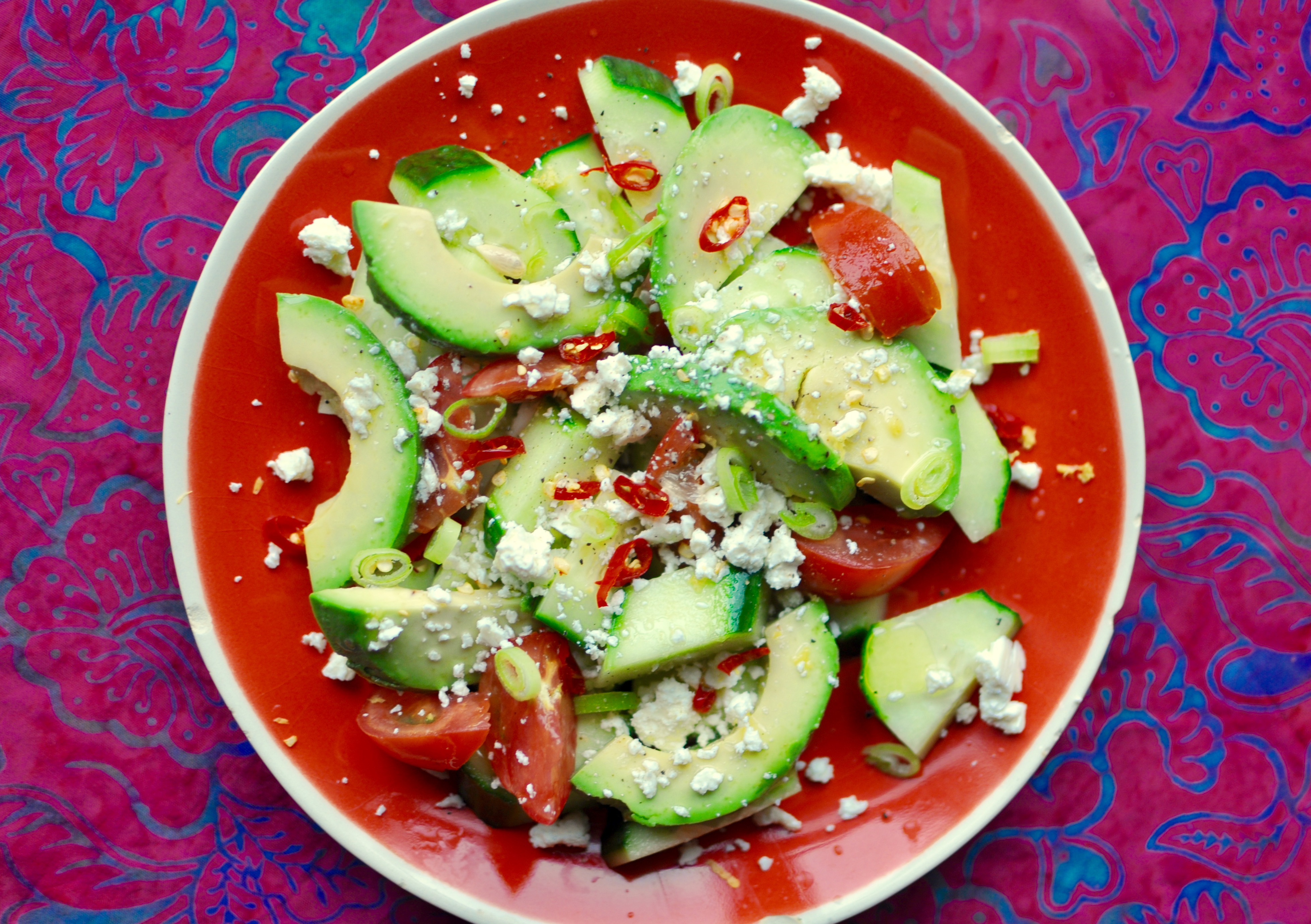An orange plate full of cucumber, tomato, and avocado salad on a bright pink and blue tablecloth. The salad is garnished with chilies, feta cheese, and scallions.