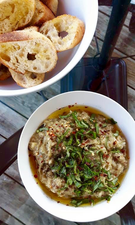 A white round bowl full of creamy eggplant dip, garnished with fresh basil and mint beside a bowl of grilled baguette slices.
