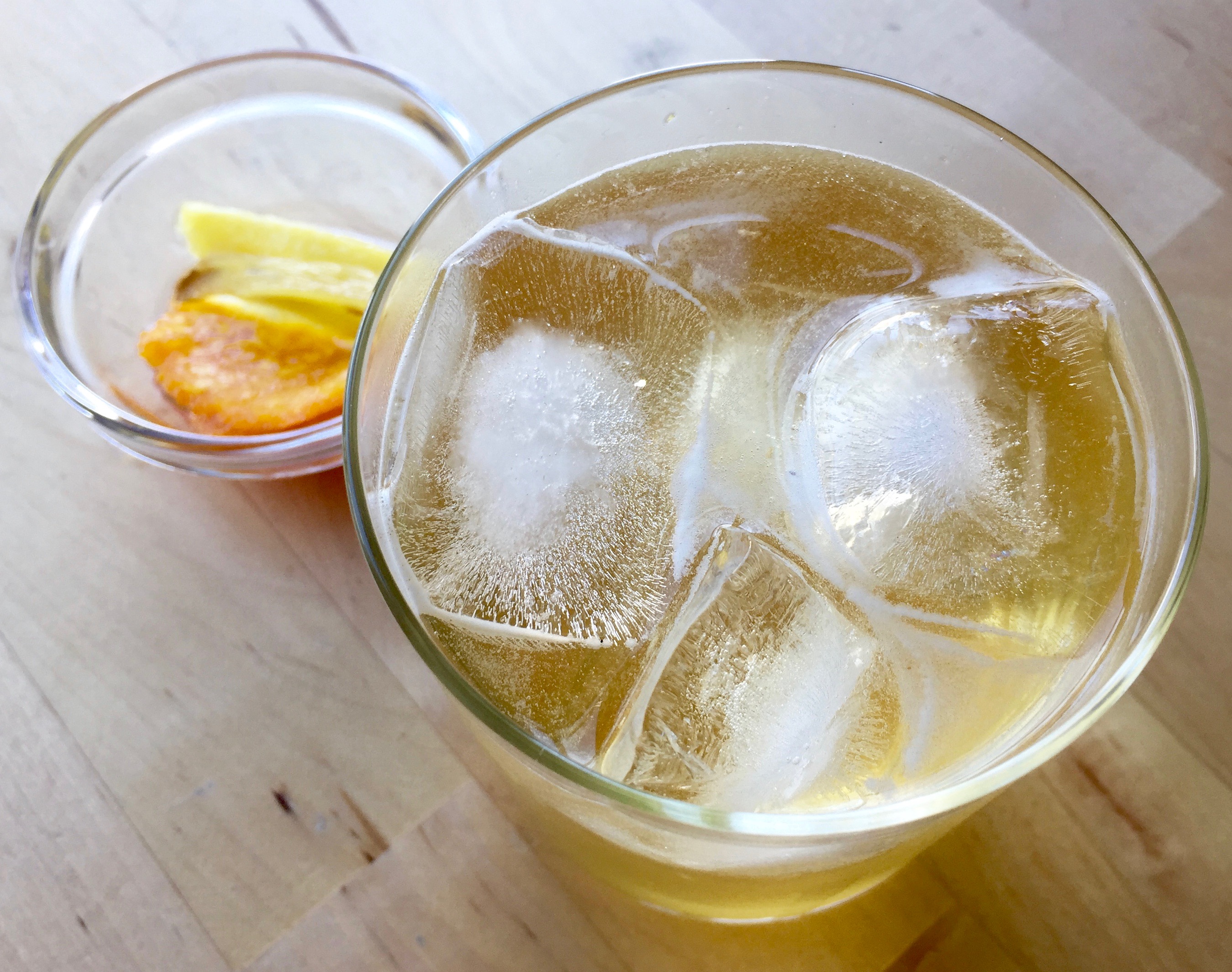 Clear glass tumbler glass containing ice, rye, ginger beer, simple syrup, and sparkling water next to a small prep bowl with candied ginger and orange peel from the simple syrup.