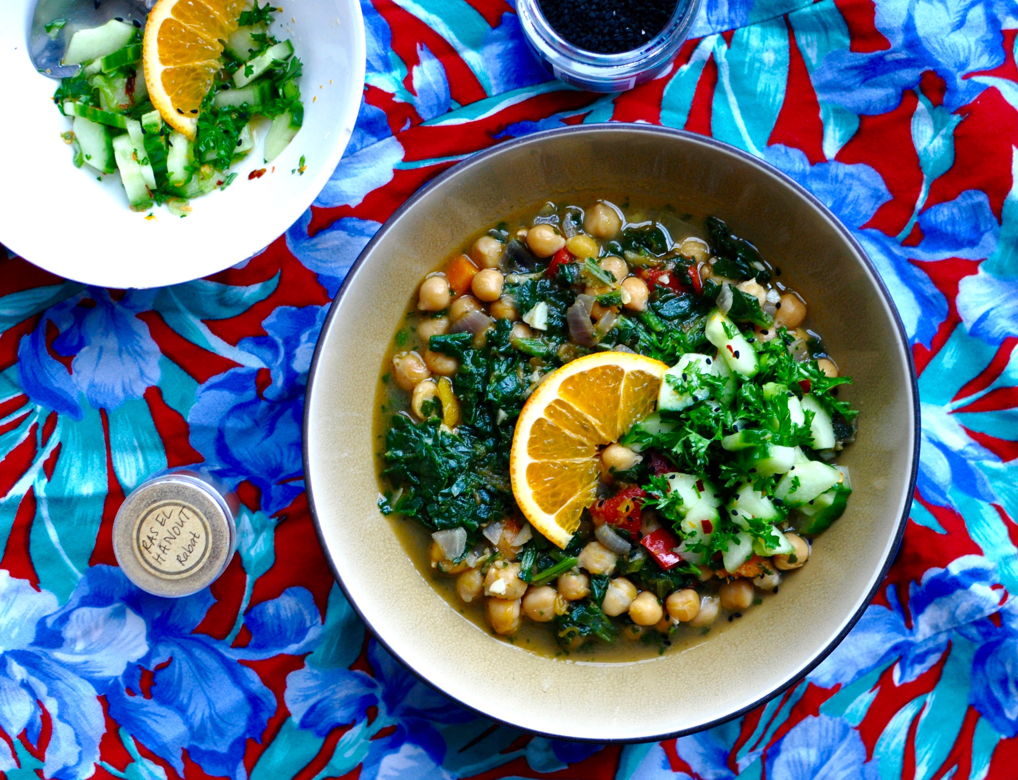 spiced chickpea stew with butternut squash and spinach in a shallow brown bowl, a small white bowl to the side holds a cucumber slaw to top the stew with. the bowls are arranged on a blue and red tablecloth, with a small pot of ras el hanout and a tub of black nigella seeds.