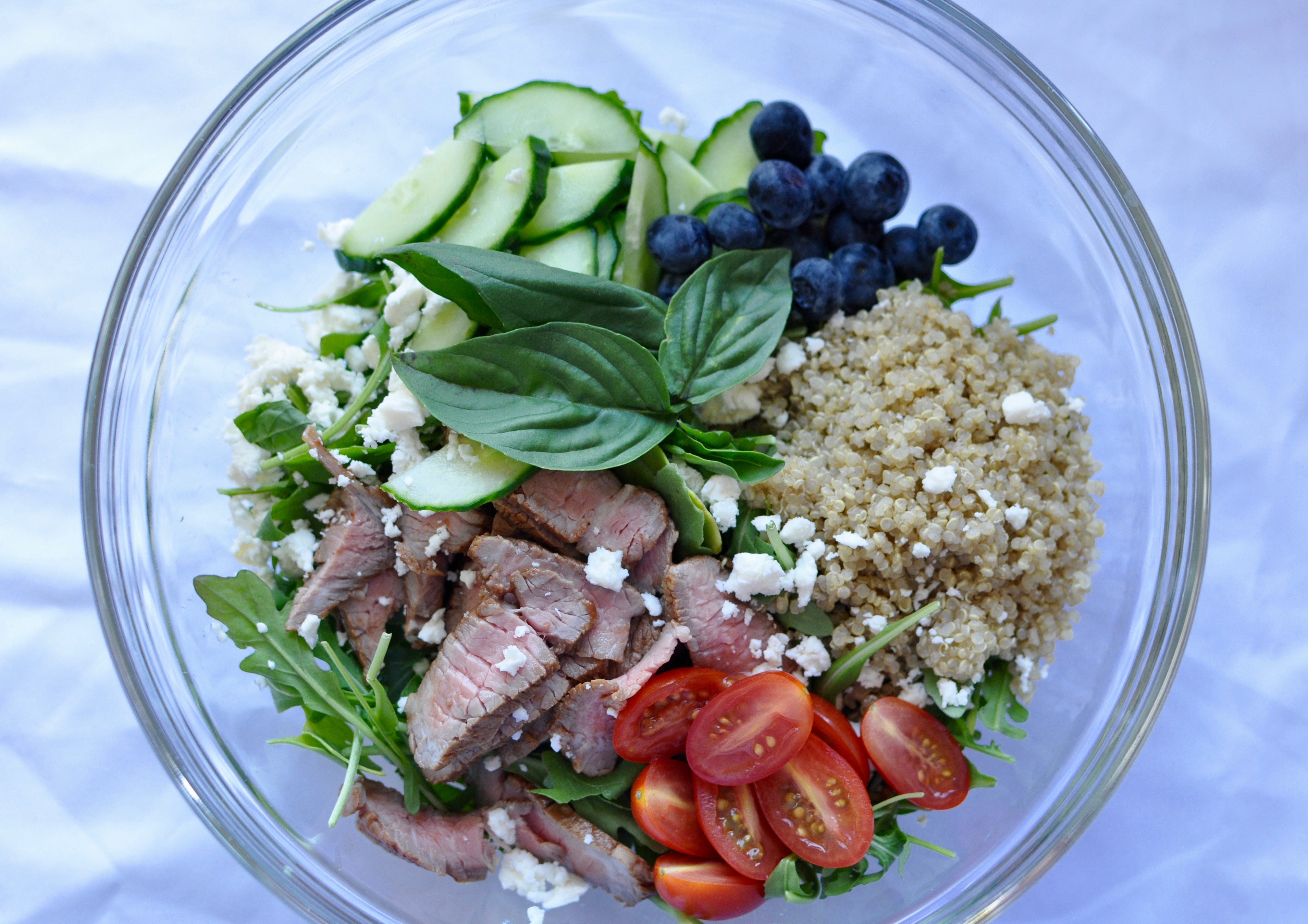 Summer salad of leftover steak, quinoa, blueberries, tomatoes, cucumber, arugula, and feta in a clear glass bowl on a white background.