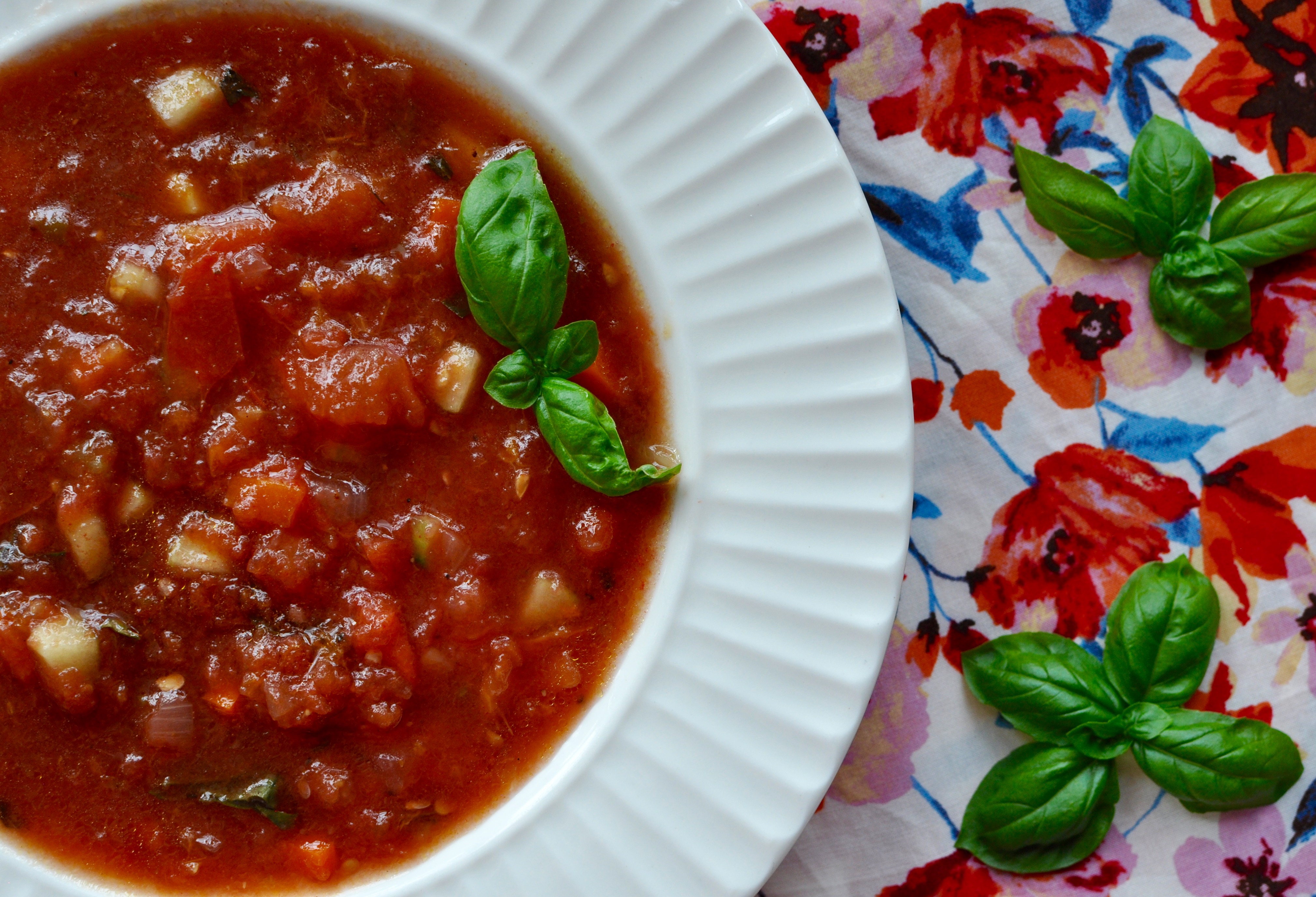 A shallow white soup bowl full of watermelon gazpacho with roasted apricots, tomatoes, and red onions on a red, white, blue floral background. Small fresh basil clusters are arranged on the gazpacho and background.