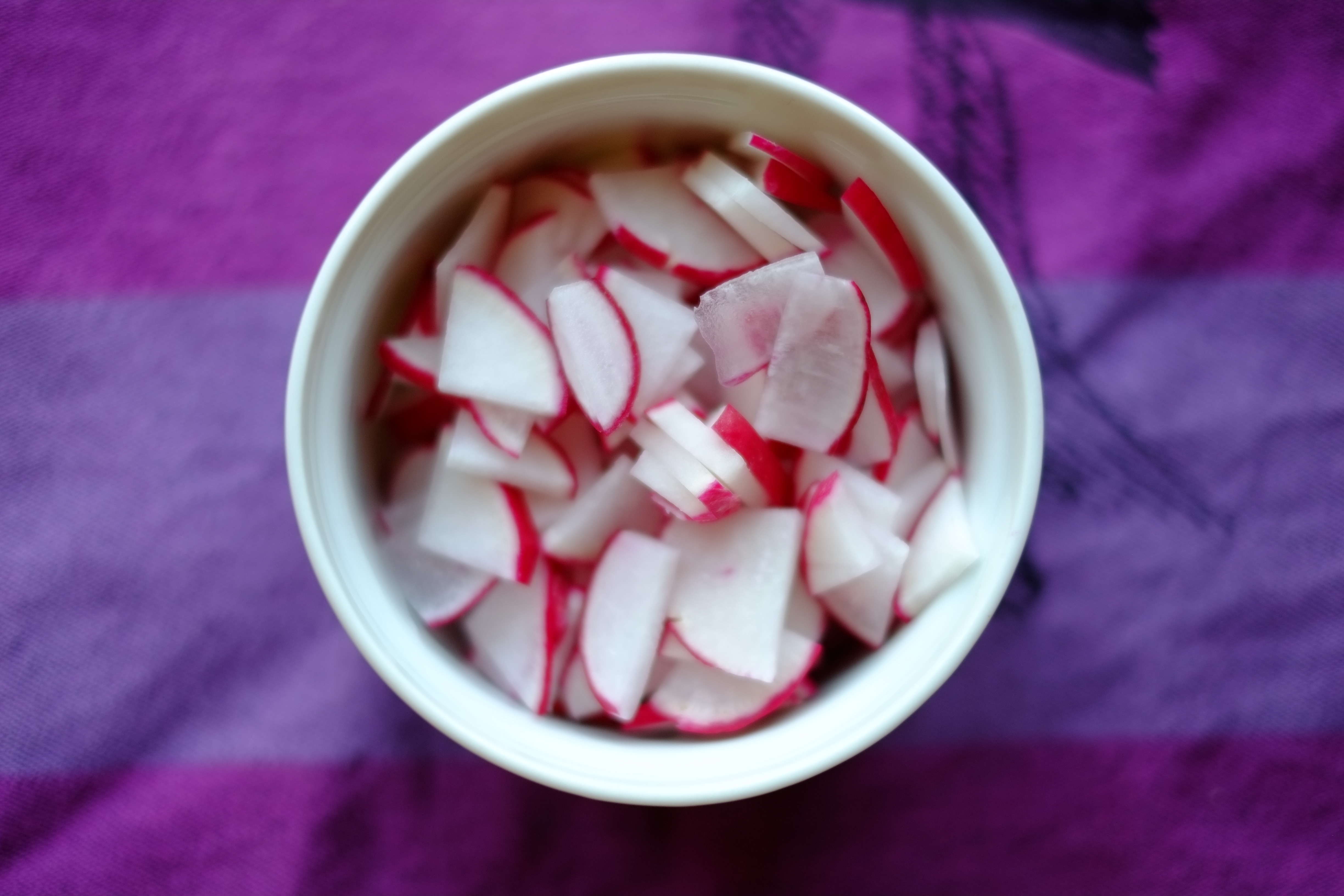 Small white ramekin with thinly sliced red radishes on a purple background.