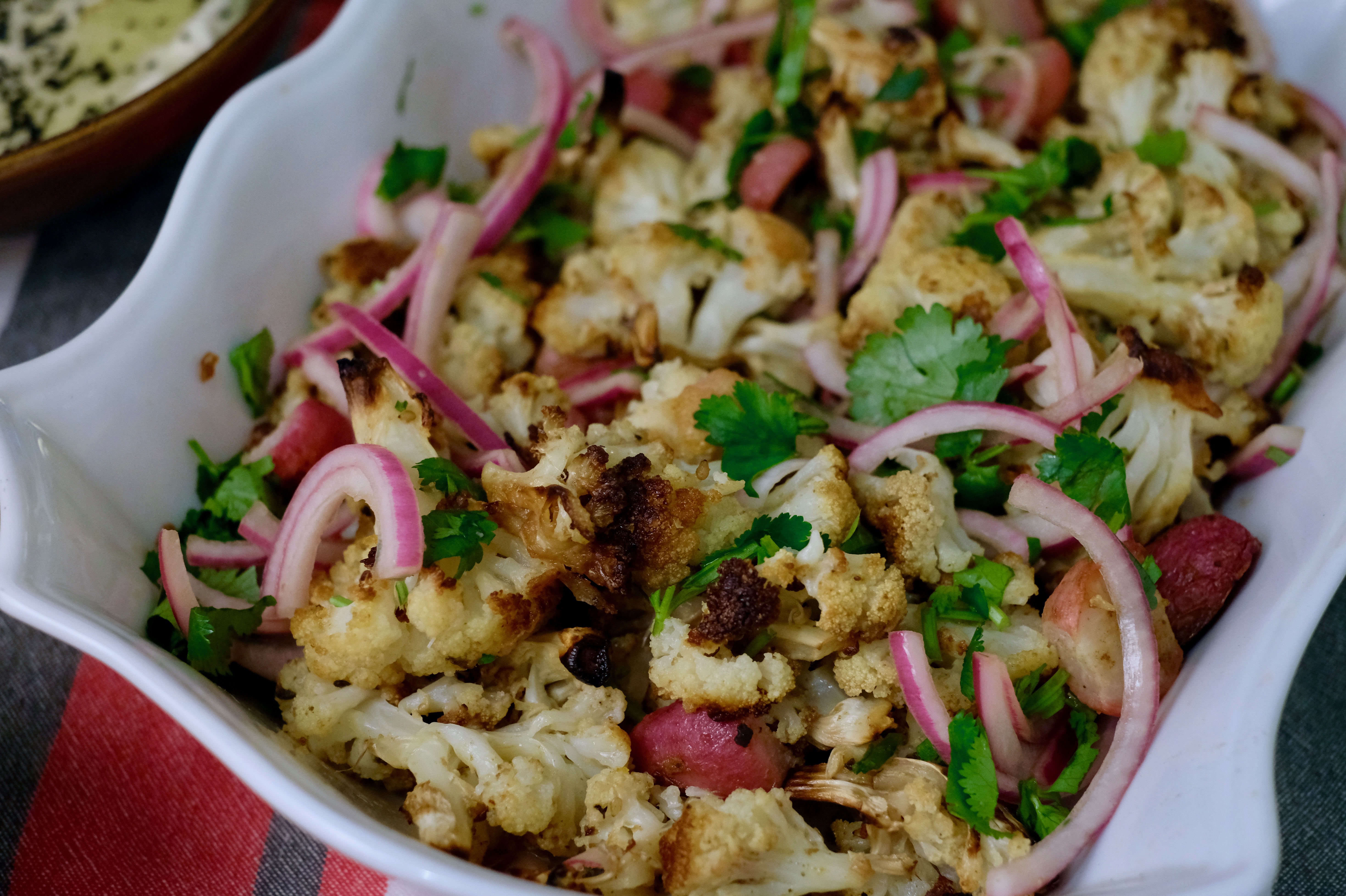 A white casserole dish full of roasted cauliflower, radishes, pickled onions, and cilantro on a red and green striped tablecloth.