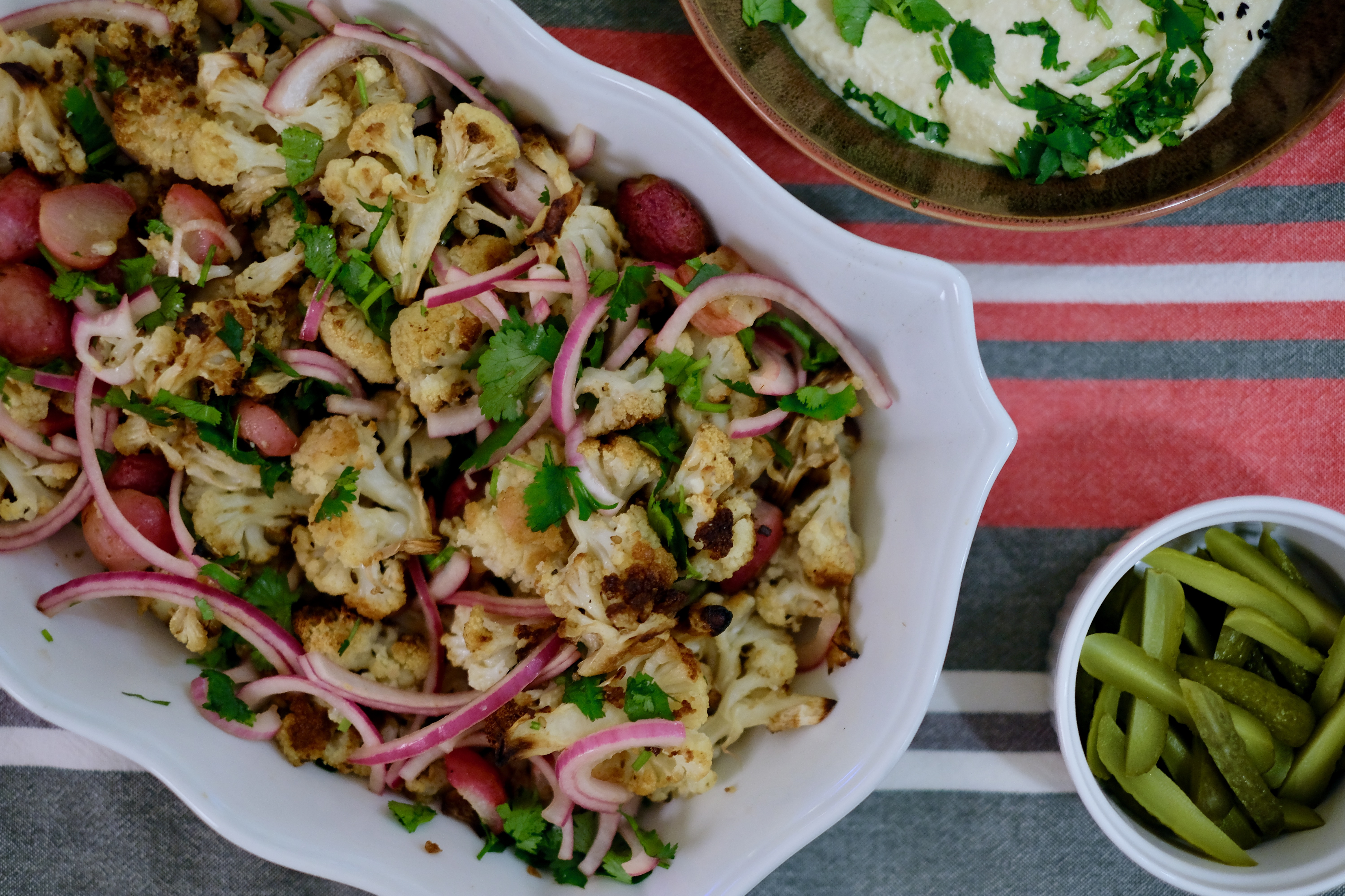 A white casserole dish full of roasted cauliflower, radishes, pickled onions, and cilantro on a red and green striped tablecloth.