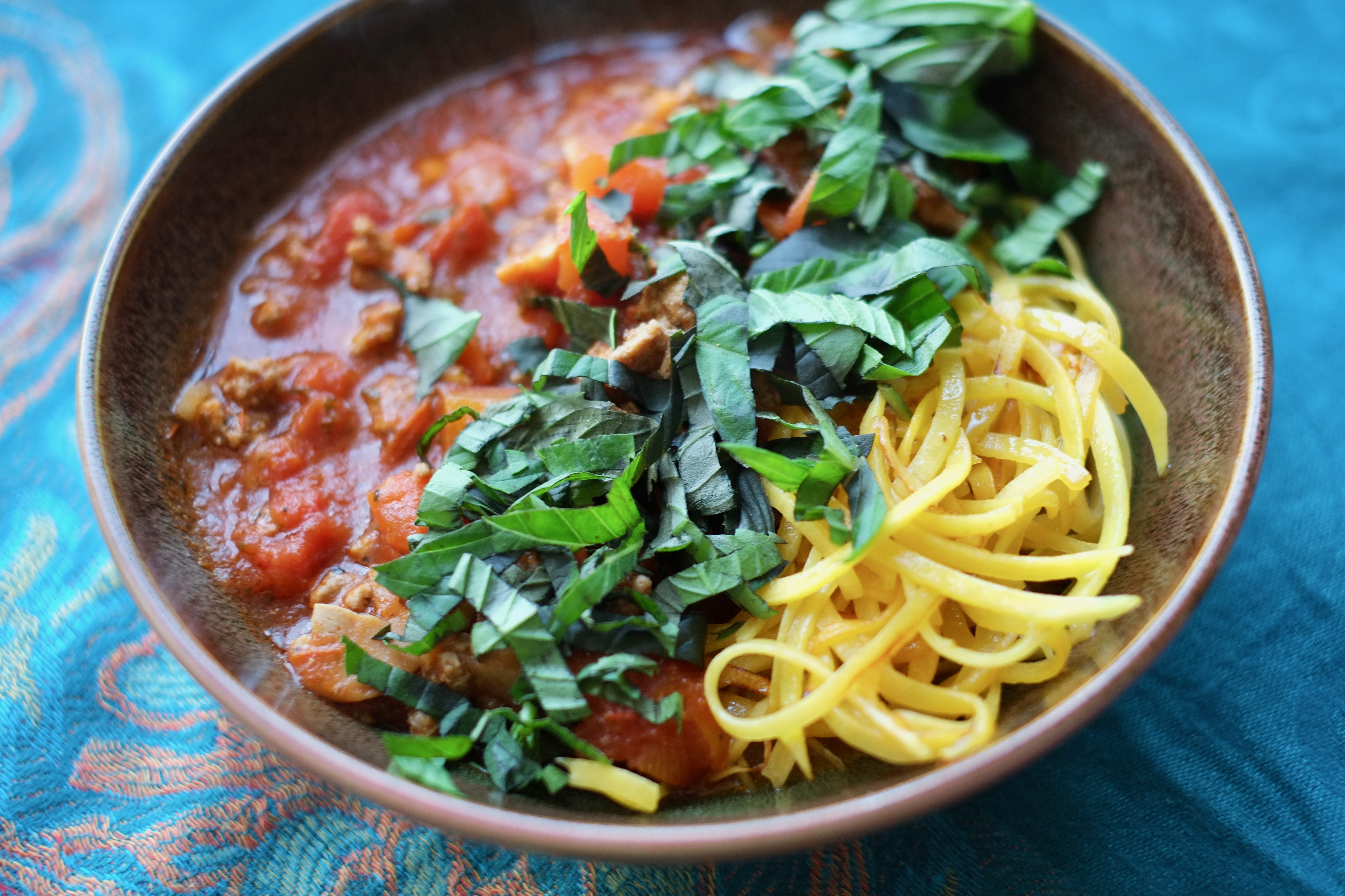 Shallow earth-coloured bowl on a turquoise tablecloth. The bowl contains a tomato and ground meat sauce, butternut squash "noodles" and shredded fresh basil.