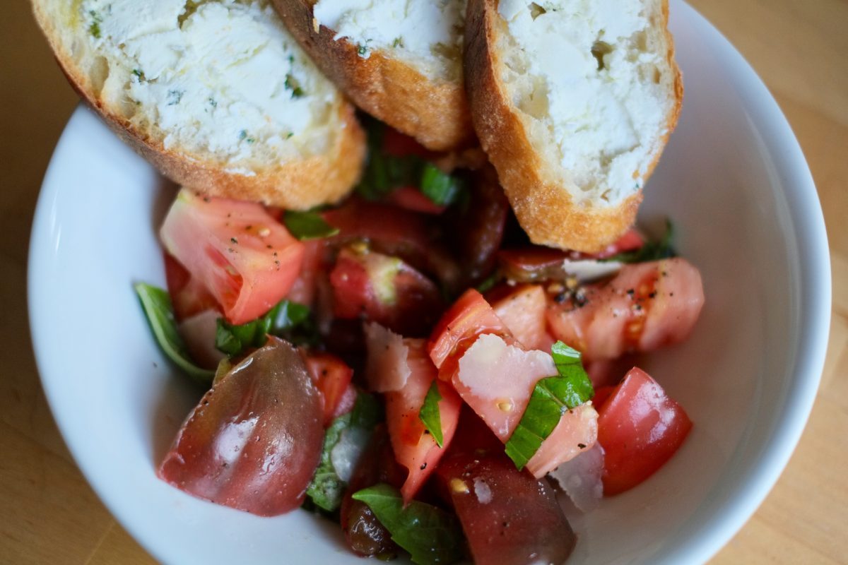 Composed summer tomato salad with fresh basil and shaved parmesan in a white shallow bowl with baguette slices and goat cheese.