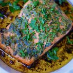 A round white baking dish that contains a fillet of baked trout in olive oil with herbs.