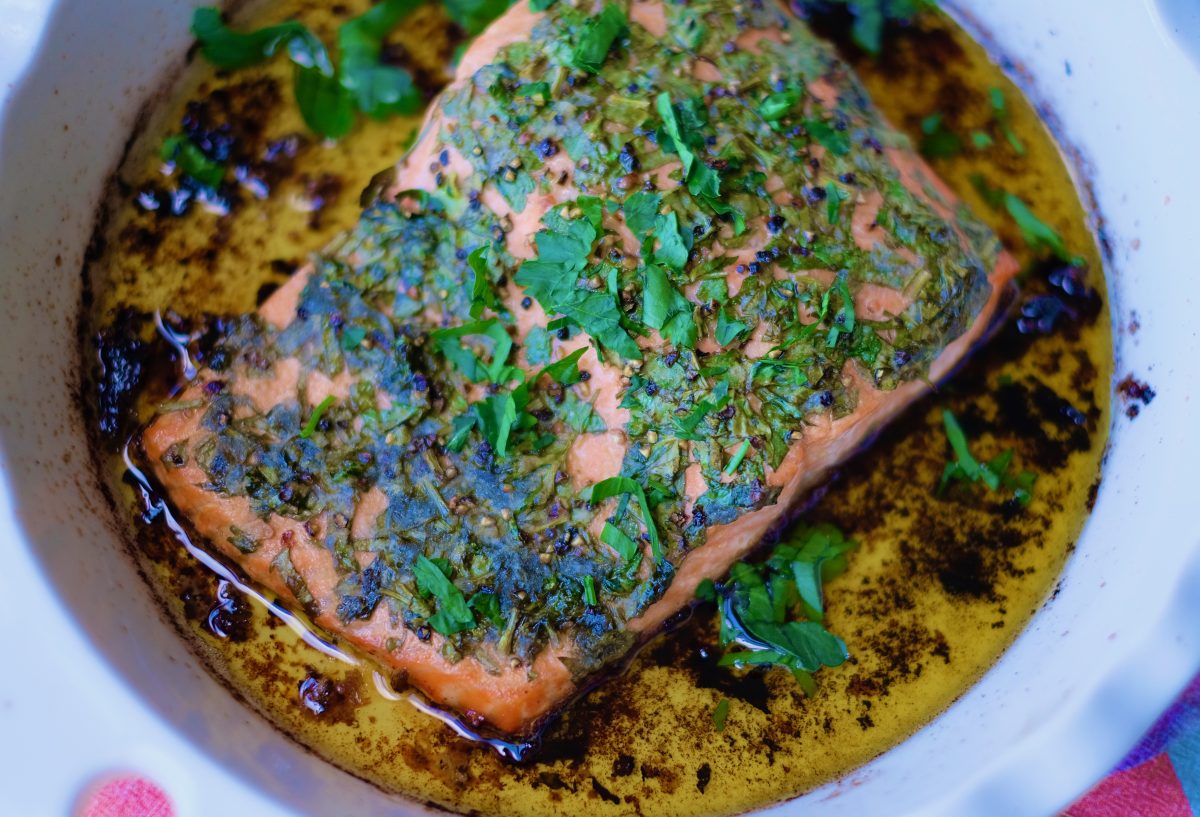 A round white baking dish that contains a fillet of baked trout in olive oil with herbs.